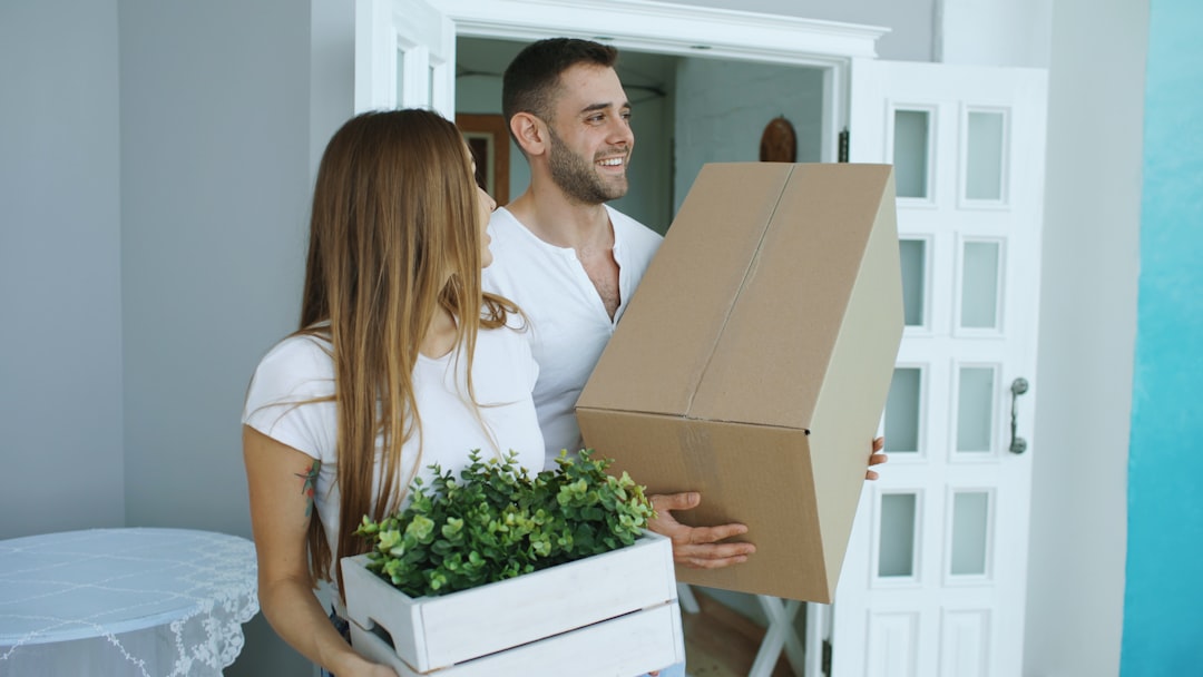 A couple carrying cardboard moving boxes through the doorway of a new house